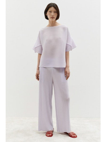 A person wears the MEIMEIJ - PLISSE TOP, a loose-fitting, light lavender lilac top with wide sleeves, against a plain white background. Their minimalist ensemble is paired with matching pleated pants, complementing their dark shoulder-length hair.