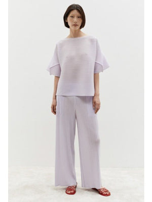 A person wears the MEIMEIJ - PLISSE TOP, a loose-fitting, light lavender lilac top with wide sleeves, against a plain white background. Their minimalist ensemble is paired with matching pleated pants, complementing their dark shoulder-length hair.