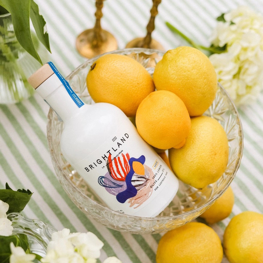 A white bottle with a wooden cap labeled &quot;BRIGHTLAND - &#39;LUCID&#39; LEMON INFUSED OLIVE OIL&quot; features a vibrant, abstract design with shapes in red, blue, yellow, and purple. The label indicates it is made from 100% lemon-infused olive oil using early-harvest Frantoio olives sourced from small family farms. The bottle holds 375 ml or 12.7 fl oz of oil.