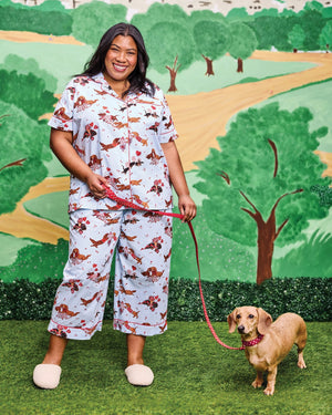 A woman with long, wavy brown hair stands barefoot against a white wall, wearing the PRINTFRESH Dachshund Date Short Sleeve Top & Cropped Pants Pajama Set in Blue. The organic cotton set from PRINTFRESH features short sleeves and cropped pants adorned with a charming dachshund print. She smiles slightly.