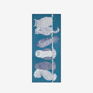 A blonde woman in a black coat stands against a white background, wearing the INOUI EDITIONS "Owls on a Branch" wool scarf in blue by INOUI, draped over her neck and one shoulder with intricate white owl designs.