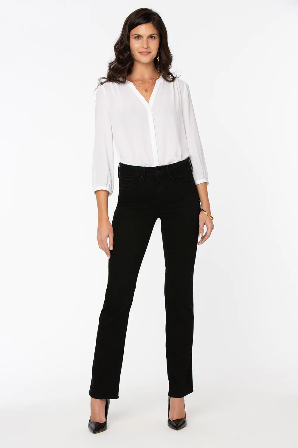 A woman with long dark hair is standing against a plain white background, wearing a white long-sleeve blouse with a V-neckline, NYDJ Marilyn Straight in Black jeans featuring Lift Tuck® Technology, and black high-heeled shoes. She has a neutral expression and her arms are relaxed at her sides.