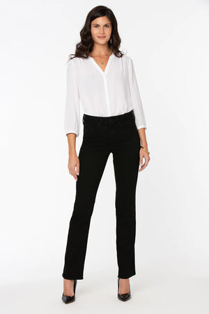 A woman with long dark hair is standing against a plain white background, wearing a white long-sleeve blouse with a V-neckline, NYDJ Marilyn Straight in Black jeans featuring Lift Tuck® Technology, and black high-heeled shoes. She has a neutral expression and her arms are relaxed at her sides.