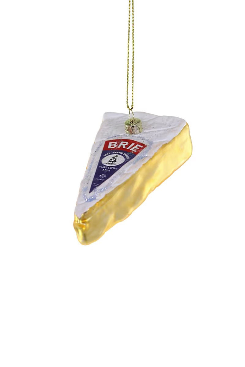 The CODY FOSTER WEDGE OF BRIE ORNAMENT is a decorative glass Christmas tree ornament shaped like a Brie wedge, with a gold edge, a label on top, and a gold string for hanging.