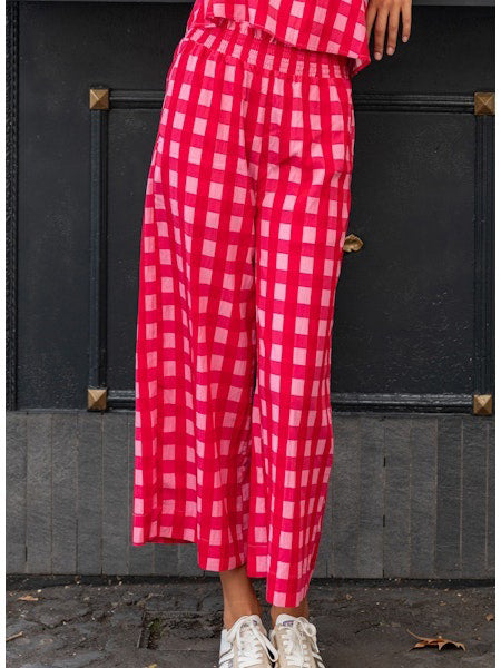 A person stands on tiled flooring wearing the MERSEA - VERANDA CROPPED PANT in navy blue with a ruffle waistband, a matching top, and red studded sandals. Their left hand shows a ring. The white wall behind them has blue accents.