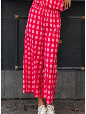 A person stands on tiled flooring wearing the MERSEA - VERANDA CROPPED PANT in navy blue with a ruffle waistband, a matching top, and red studded sandals. Their left hand shows a ring. The white wall behind them has blue accents.