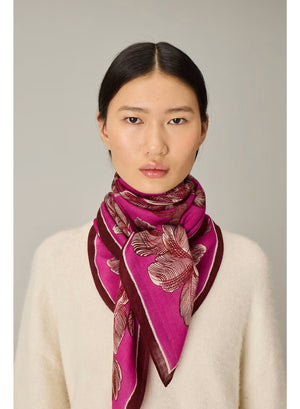 A woman with straight black hair wears a cream sweater and the INOUI EDITIONS - ARBORI Square Wool Scarf in Fuschia by INOUI, crafted from 100% wool and featuring a leaf pattern, draped around her neck against a plain light background.