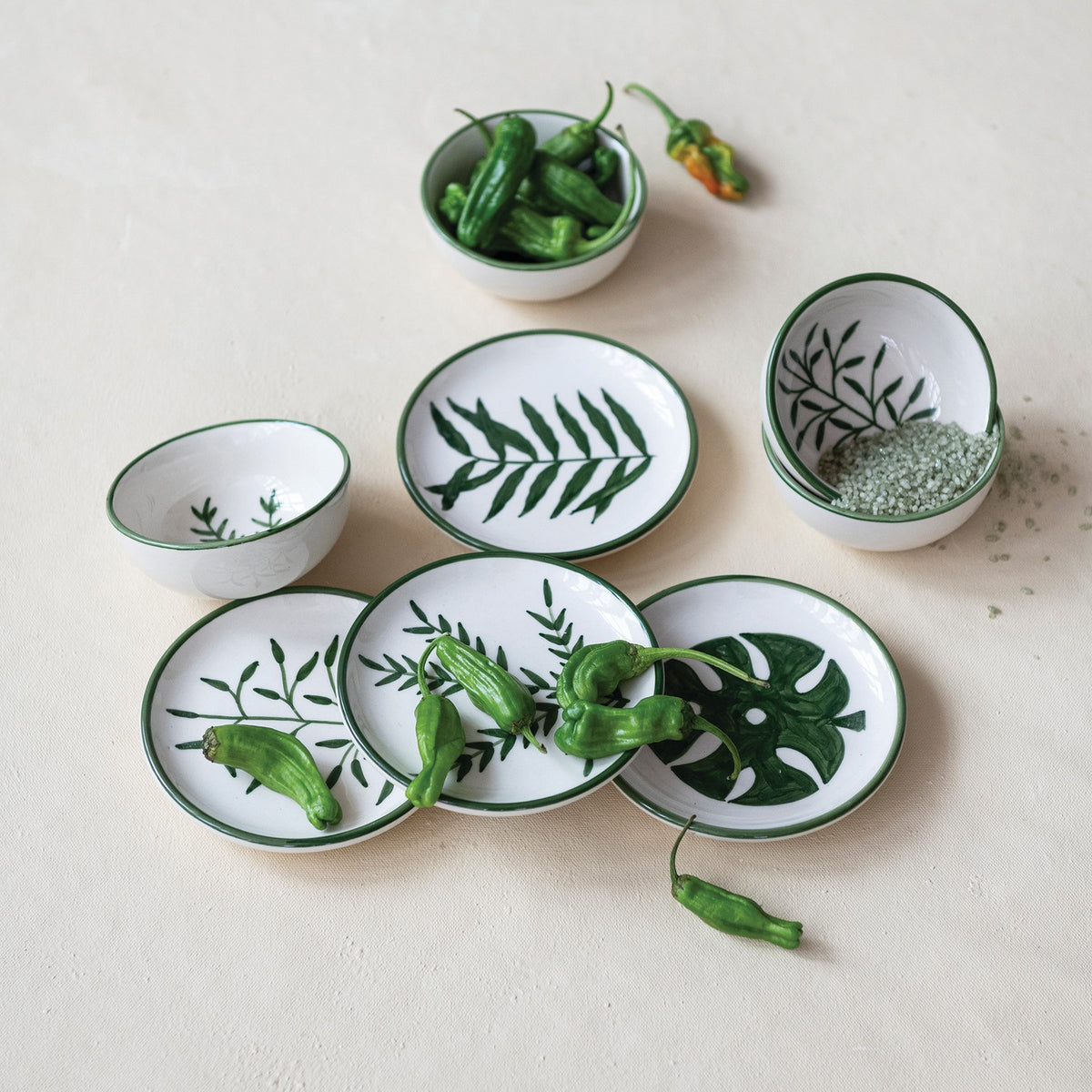 CREATIVE COOP's HAND-PAINTED STONEWARE BOWLS WITH BOTANICAL DESIGN, some holding green peppers or grains, are arranged on a light beige surface—each bowl highlights its unique handcrafted artistry.