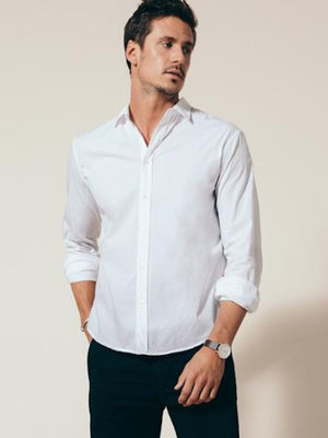 A man in the FRANK & EILEEN Finbar Men's Button Up Shirt in classic white poplin stands with rolled sleeves and hands in pockets, looking to the side. He has short dark hair, wears a watch, and poses against a light background.