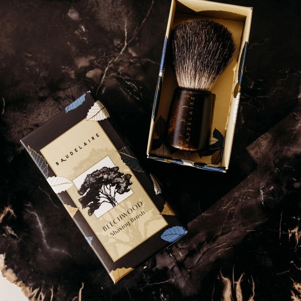 The BAUDELAIRE - M.B. SHAVING SET by BAUDELAIRE includes a luxurious shaving brush and premium shaving soap, presented in an elegant box featuring a delightful hat illustration and “shaving set” printed on the front. Displayed outdoors atop lush moss, this refined ensemble adds classic sophistication to your grooming routine.