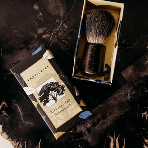 The BAUDELAIRE - M.B. SHAVING SET by BAUDELAIRE includes a luxurious shaving brush and premium shaving soap, presented in an elegant box featuring a delightful hat illustration and “shaving set” printed on the front. Displayed outdoors atop lush moss, this refined ensemble adds classic sophistication to your grooming routine.