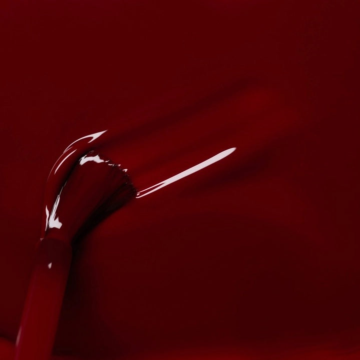 A hand with neatly manicured nails painted in a rich shade of dark red rests on red fabric beside a matching bottle of MANUCURIST INC’s GREEN™ Nail Polish in Dark Pansy, a non-toxic and vegan formula.