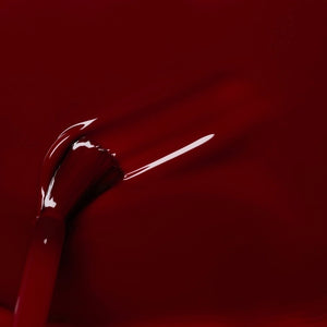 A hand with neatly manicured nails painted in a rich shade of dark red rests on red fabric beside a matching bottle of MANUCURIST INC’s GREEN™ Nail Polish in Dark Pansy, a non-toxic and vegan formula.