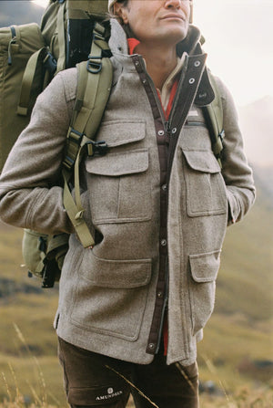 A man wearing the AMUNDSEN SPORTS - HUNTSMAN JACKET over a cream turtleneck sweater and tan pants stands facing the camera against a white background.