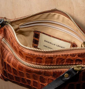 The DANIELLA LEHAVI - LORRIE CLUTCH, a small brown leather wristlet with a crocodile pattern, is displayed on a white pedestal against a light wooden background, highlighting its sophisticated Italian craftsmanship.