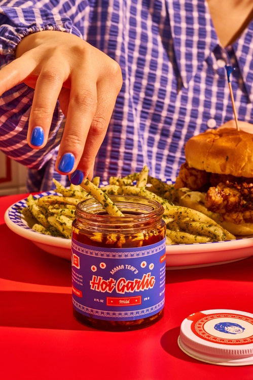 A jar of MAMA TEAV'S- MILD HOT GARLIC CHILI CRISPS is displayed against a plain white background. The jar features a blue label with the brand name "MAMA TEAV'S" prominently in the center and the word "MILD" beneath it, describing the product inside. Made with natural ingredients, this gluten-free Hot Garlic Chili Crisp is perfect for any meal.