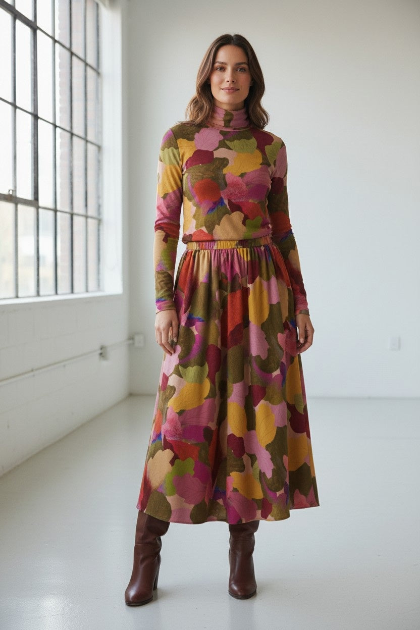A person stands indoors near sunlight, dressed in a long-sleeved top paired with the FRANCO FERRARI - FRAGOLINA SKIRT by FRANCO FERRARI, which features a bold and colorful floral pattern in shades of pink, red, green, and yellow.