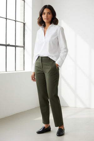 A woman with long blonde hair models the HINSON WU Monica Long Sleeve Button Back Top in white—a collared stretch polished cotton blouse featuring a split neckline and long sleeves—paired with dark green pants, standing against a plain light background.