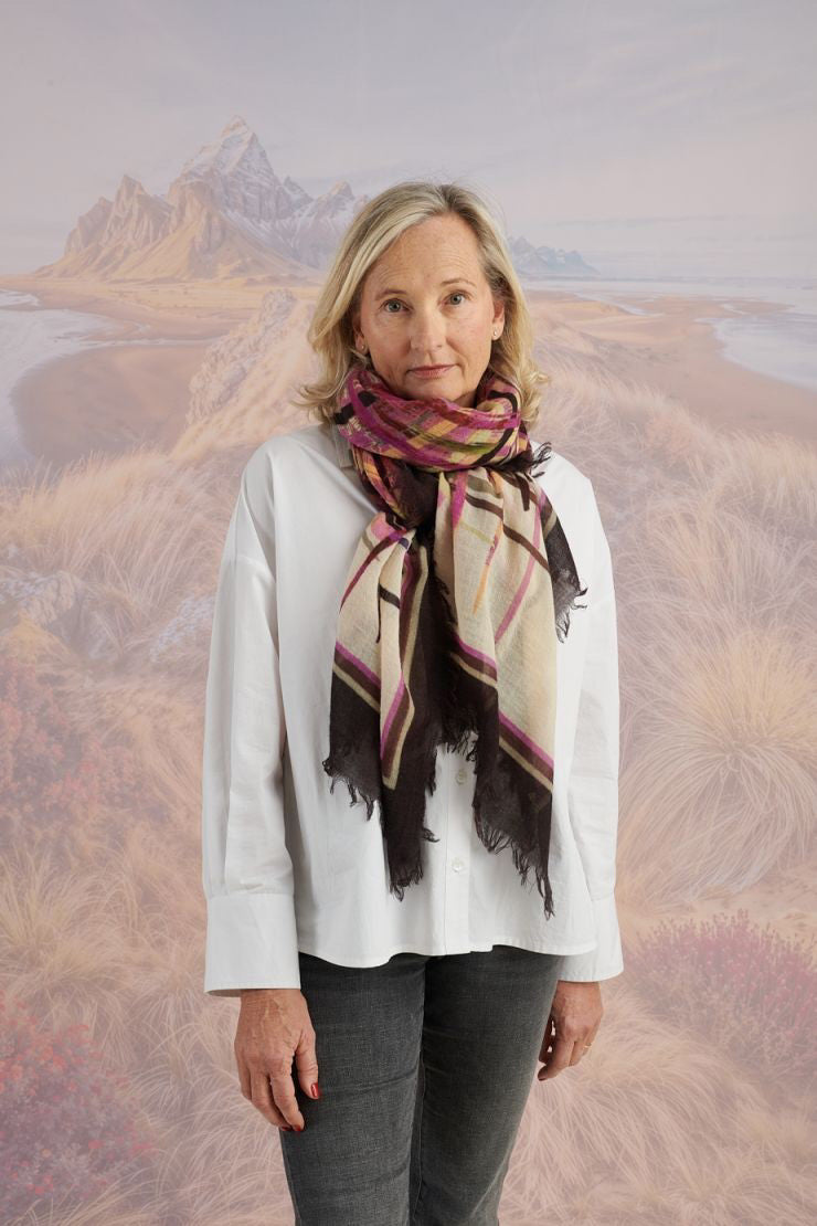 A woman in a blue shirt and jeans stands holding the STORIATIPIC - JULIE WOOL SCARF, a luxury plaid accessory in earthy tones. Behind her, mountains, beach, and grassy dunes create a scenic backdrop.