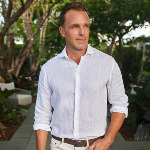 Wearing a COLLARS AND CO - TOSCANO LINEN SHIRT IN WHITE, a man with sunglasses sits on a red scooter by a scenic coastal road, holding a white helmet, with hills, buildings, and the sea in the background.
