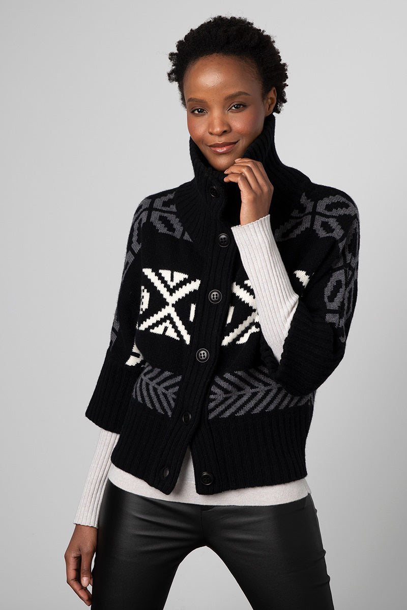 A person is wearing the KINROSS CASHMERE - CROP NORDIC COWL CARDIGAN by KINROSS, featuring a cozy black and gray pattern crafted from luxurious wool. The cardigan has a high collar and button front. They strike a relaxed, confident pose with one hand touching their face, paired with a light gray undershirt and black pants.