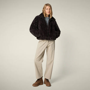 A woman models the SAVE THE DUCK - ELINA REVERSIBLE JACKET in beige fur, styled with a white skirt, orange tights, burgundy boots, and a matching clutch, standing against a plain light background.