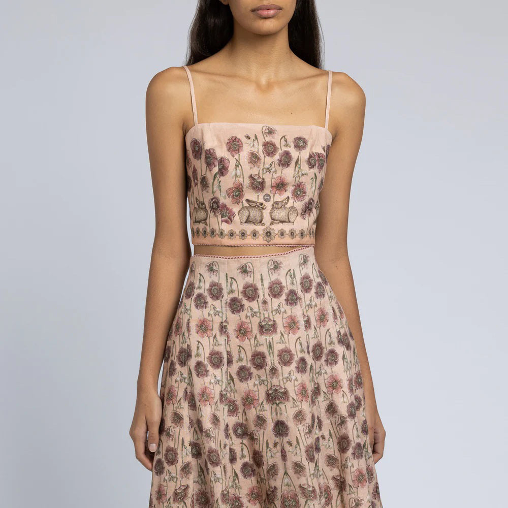 A woman is wearing a light pink floral two-piece outfit. The SABINA SAVAGE - HONOURING ARGOS CROPPED FITTED TOP from SABINA SAVAGE ENGLAND is sleeveless with thin straps and a square neckline, paired with a high-waisted skirt that flows gracefully from the hips. Crafted from pure linen, the fabric features a pattern of small, detailed floral designs perfect for any Greek-inspired look.