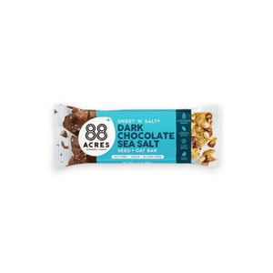 An "88 ACRES - DARK CHOCOLATE SEA SALT SEED OAT BAR" in blue and white packaging rests on a white wooden surface. The packaging reads "Sweet 'n' Salty Dark Chocolate Sea Salt Seed + Oat Bar." This 1.7-ounce bar, from the brand 88 ACRES, is nut-free, vegan, gluten-free, and packed with flavors reminiscent of chocolate chip cookies.