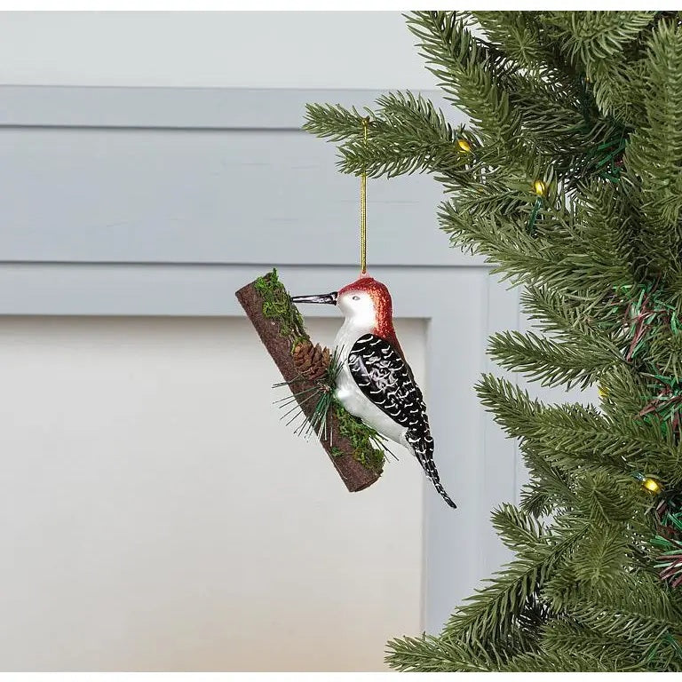 WOODPECKER ON TREE ORNAMENT featuring a glass woodpecker perched on a decorative branch with small pinecones and moss, perfect for woodland holiday decor.