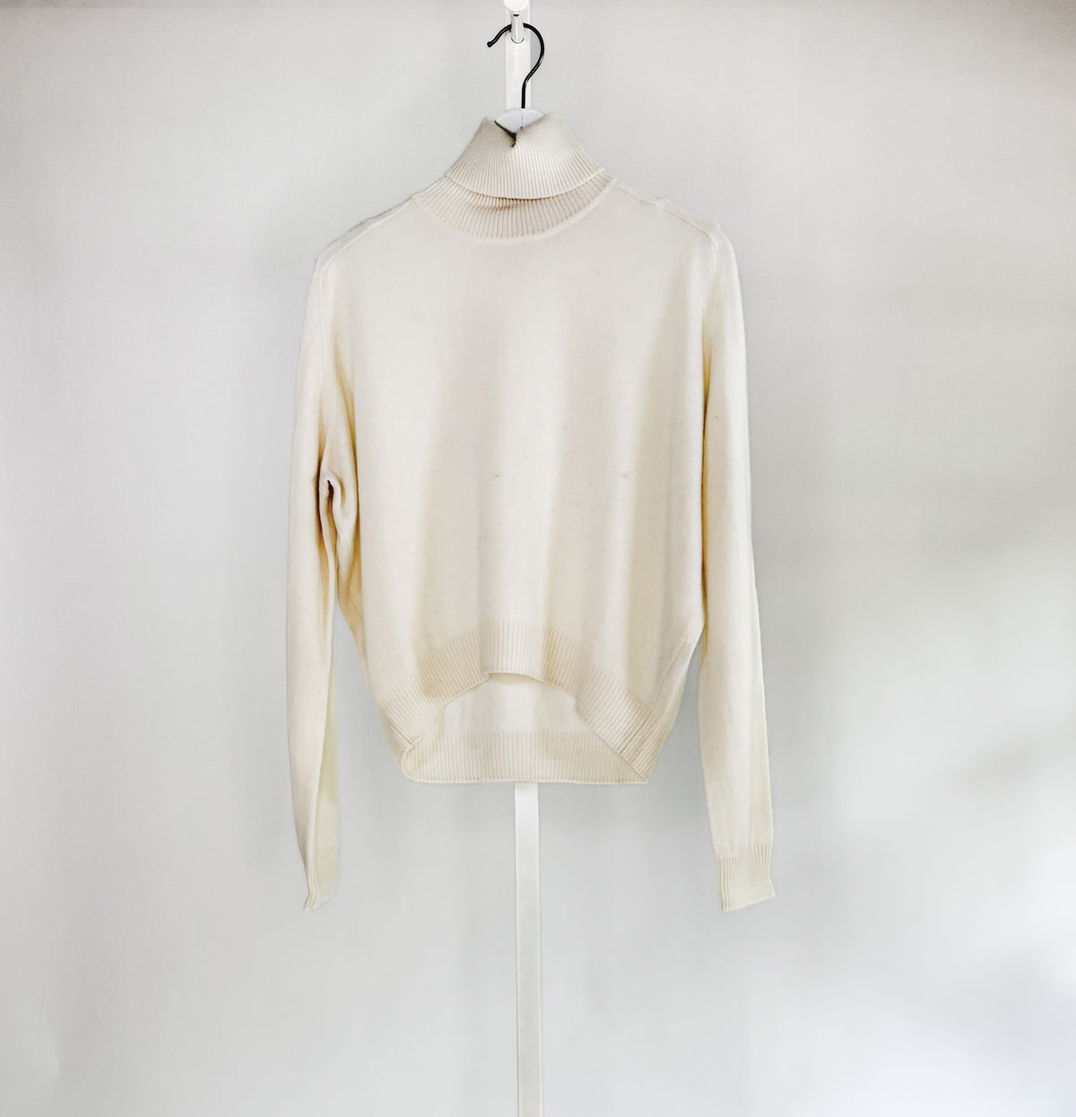 A woman with long brown hair smiles against a white background, wearing a ROSE &amp; CROWN - CASHMERE TURTLENECK and wide-legged green pants, accessorized with necklaces, bracelets, and a large black handbag.