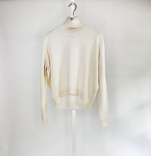 A woman with long brown hair smiles against a white background, wearing a ROSE & CROWN - CASHMERE TURTLENECK and wide-legged green pants, accessorized with necklaces, bracelets, and a large black handbag.