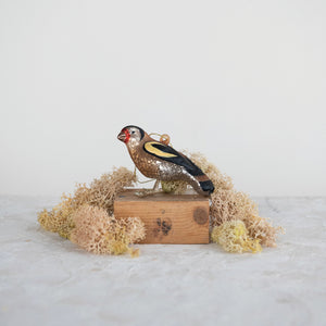 The CREATIVE COOP BIRD ORNAMENT WITH GLITTER features realistic brown, yellow, and black coloring with a shimmering red face patch, detailed feet, and hangs by a gold string against a white background.