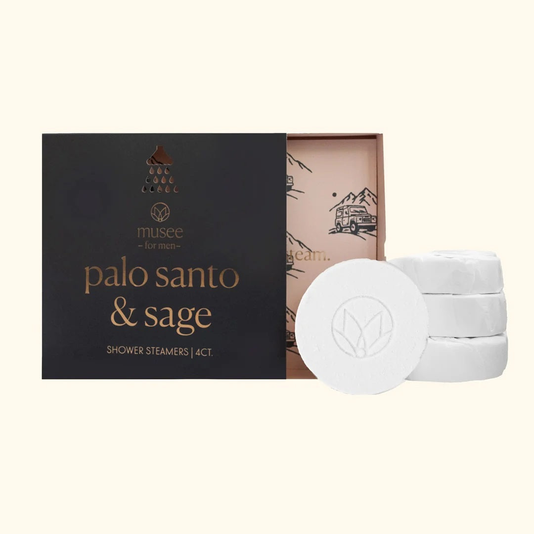 A person wearing a blue robe holds a black box labeled “MUSEE – Palo Santo & Sage Shower Steamers.” Only their hands and torso are visible as they display the 4-count aromatherapy set, which blends soothing palo santo and sage fragrances for a refreshing shower, from the brand MUSEE.