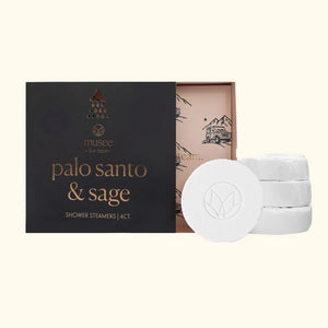 A person wearing a blue robe holds a black box labeled “MUSEE – Palo Santo & Sage Shower Steamers.” Only their hands and torso are visible as they display the 4-count aromatherapy set, which blends soothing palo santo and sage fragrances for a refreshing shower, from the brand MUSEE.