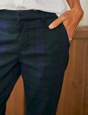 A person wearing FRANK & EILEEN's Wicklow Italian Chino in Black Watch Plaid Performance Twill, a white rolled-sleeve shirt, and black heels stands indoors, facing away from the camera with a wooden sideboard behind them.