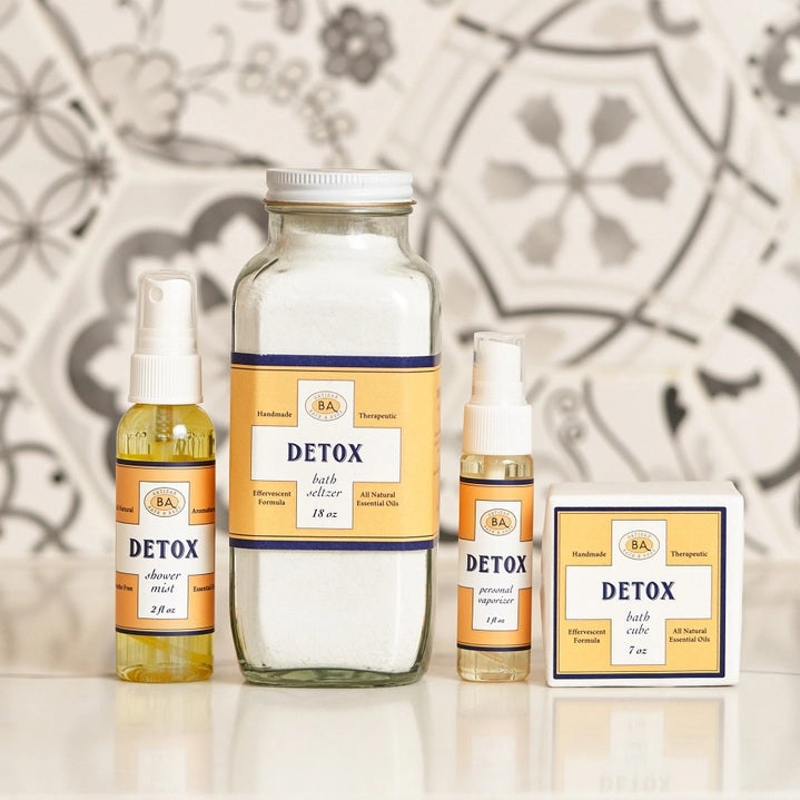 Six small bottles labeled Breathe, Boost, Unwind, Sleep, Detox, and Recover—each filled with essential oil shower mists like uplifting lemon and ginger—are arranged on white shelves in a tiled shower niche with a geometric gray and white pattern. Create your own aromatherapy steam room right at home.