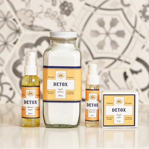 Six small bottles labeled Breathe, Boost, Unwind, Sleep, Detox, and Recover—each filled with essential oil shower mists like uplifting lemon and ginger—are arranged on white shelves in a tiled shower niche with a geometric gray and white pattern. Create your own aromatherapy steam room right at home.