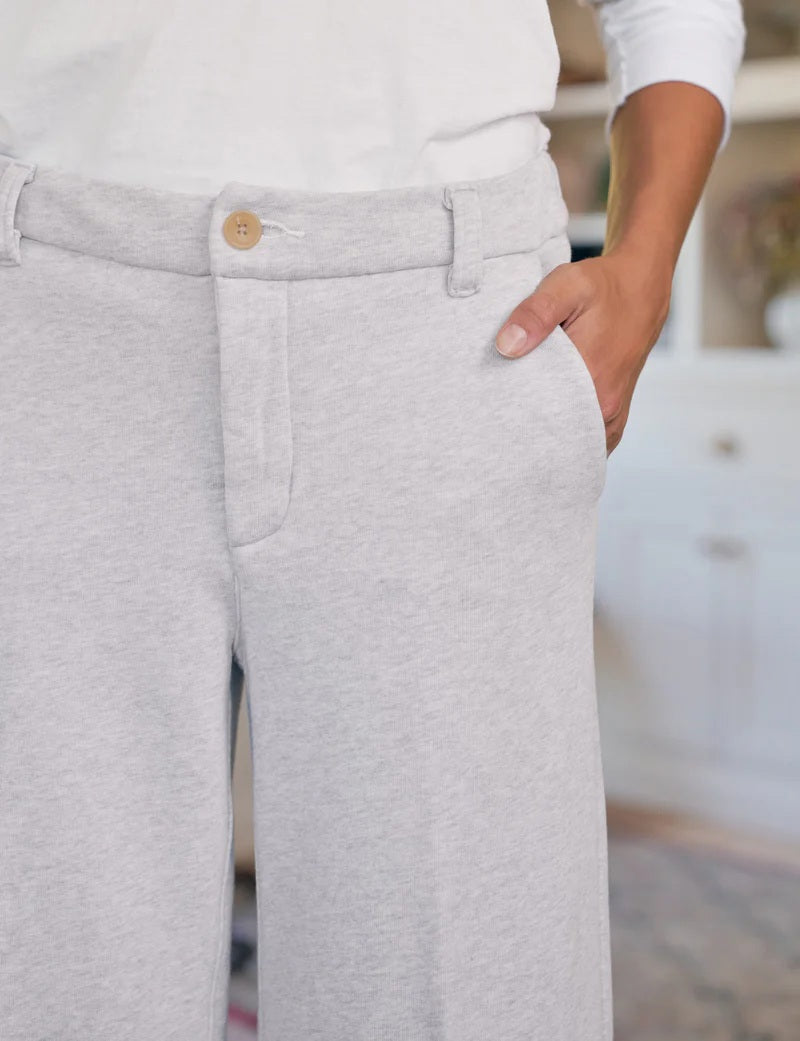 Indoors, a person wears FRANK & EILEEN's RILEY Wide Leg Trouser Sweatpant in light gray with a button waistband and a white long-sleeve top, one hand in their pocket. A red desk, wooden floor, and white cabinets complete the California Heritage Collection scene.
