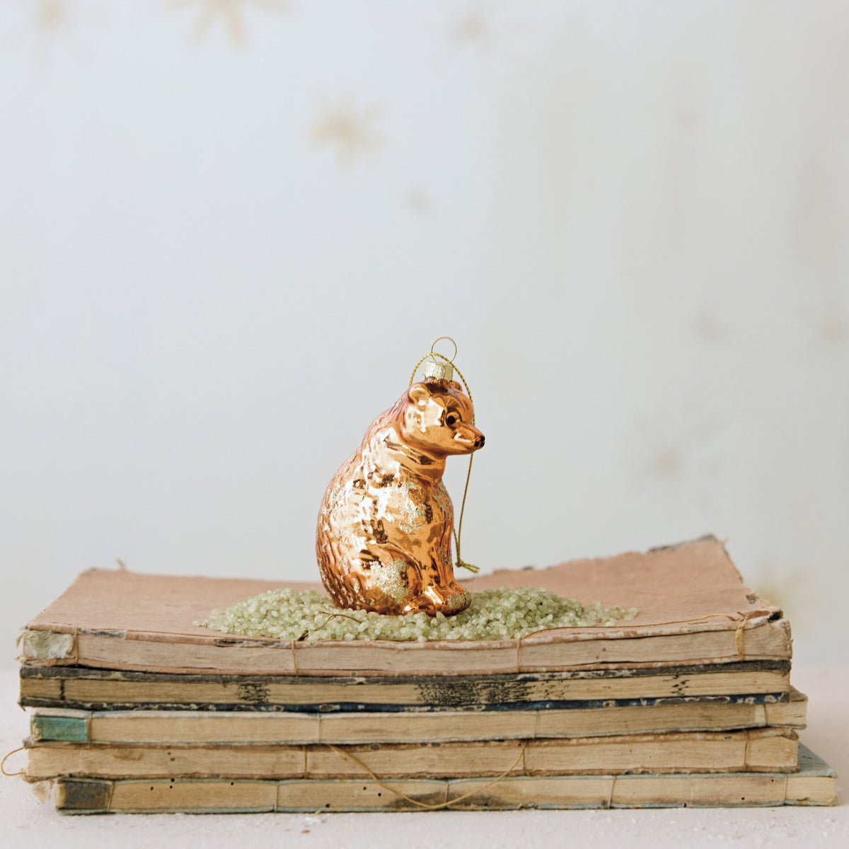 A metallic gold mercury glass BEAR ORNAMENT WITH GLITTER sits on a small pile of green glitter atop a stack of old books.