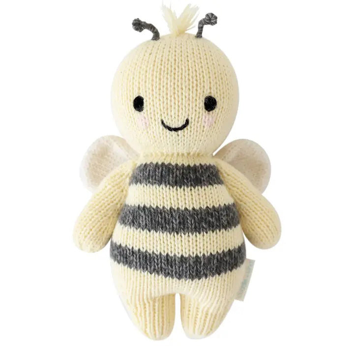 A baby lies on a cozy white blanket, raising both arms. The baby is dressed in a peach outfit and a headband with three pom-poms. Next to the baby is the CUDDLE + KIND - HANDMADE BABY BEE by CUDDLE &amp; KIND, a fair trade, hand-knit toy with black and white stripes—a perfect addition to any collection of baby shower gifts.