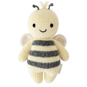 A baby lies on a cozy white blanket, raising both arms. The baby is dressed in a peach outfit and a headband with three pom-poms. Next to the baby is the CUDDLE + KIND - HANDMADE BABY BEE by CUDDLE & KIND, a fair trade, hand-knit toy with black and white stripes—a perfect addition to any collection of baby shower gifts.