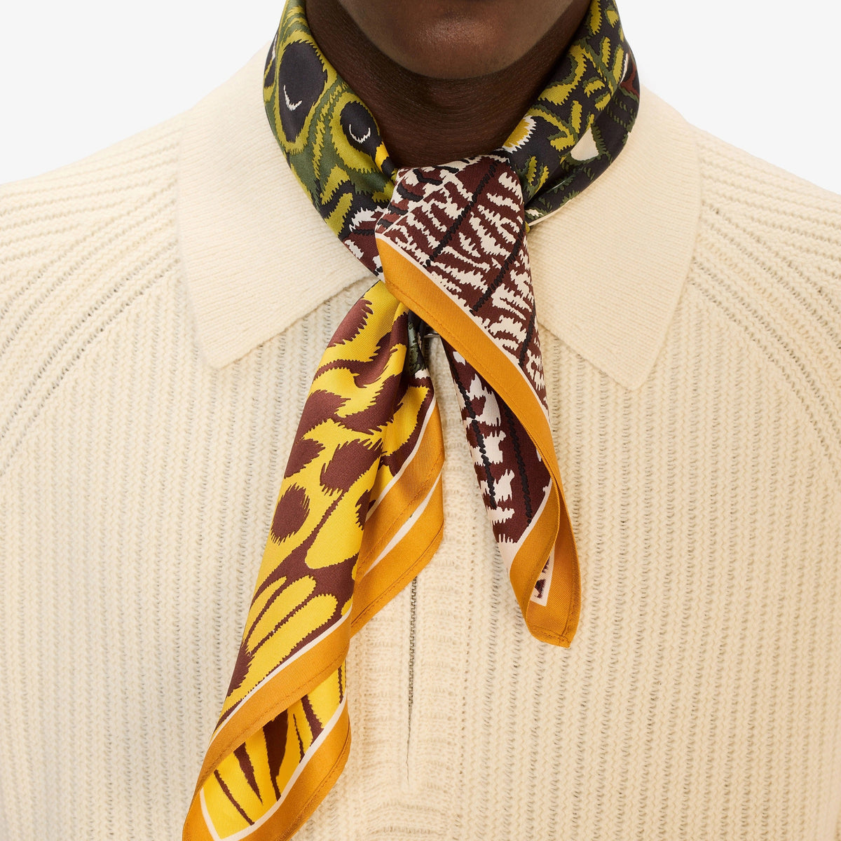 A close-up of a woman wearing a beige blazer and the INOUI EDITIONS - PAPILLON (BUTTERFLIES) Silk Scarf in Green, featuring abstract patterns in yellow, black, green, and white. Only her lower face and neck are visible.