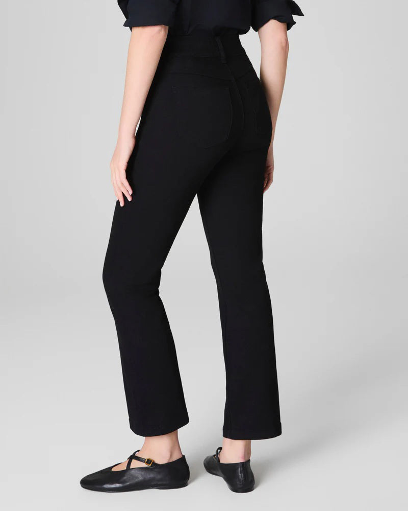 A woman stands against a light gray background, smiling with her hands behind her back. She wears the SPANX KICK FLARE JEAN IN CLASSIC BLACK, a high-waisted jean made from 4-way stretch denim, paired with a black button-up shirt and black flats.