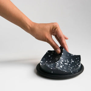 UME STUDIO’s UME - DAILY DETOX ERODE SOAP, a small round black soap with white splatter patterns, is displayed in a square white gift box with black padding. The lid and an extra box with a circular cutout sit beside it on a white surface.