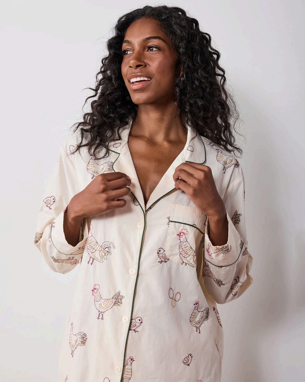 A woman dressed in the PRINTFRESH Embroidered Hens Long Pajama Set in Buff smiles as she stands by a wooden fence, holding a basket and watching chickens peck at the ground in a farmyard setting, with trees and a path visible behind her.