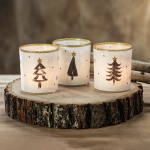 Three Zodax TREE TEALIGHT HOLDER cylindrical glass candle holders, sold individually, decorated with hand-painted black pine trees, gold glitter stars, and gold rims in an assorted style.