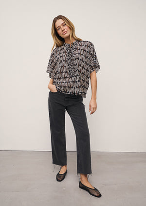 A woman stands against a light wall, wearing the DAISY BLOUSE IN BLACK AND WHITE PRINT by 0039 ITALY, black wide-leg jeans with frayed hems, and black mesh flats. One hand is in her pocket and she looks relaxed.