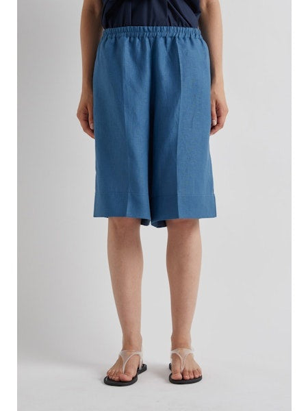 A woman stands against a plain background wearing the MEIMEIJ Pull On Linen Short in Cerulean, styled with a sleeveless navy wrap top, flat sandals, and her hair pulled back. Her expression is neutral.