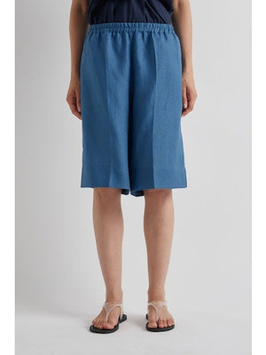 A woman stands against a plain background wearing the MEIMEIJ Pull On Linen Short in Cerulean, styled with a sleeveless navy wrap top, flat sandals, and her hair pulled back. Her expression is neutral.