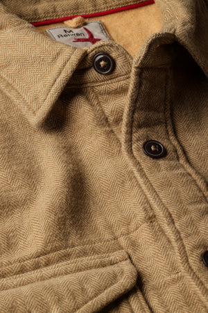 Dressed in the RELWEN Utility Workshirt in cornmeal herringbone layered over a blue and white striped shirt and paired with green pants, the individual stands with relaxed hands by their sides, capturing the effortless sophistication of luxury workwear. The person’s head is partially out of frame.